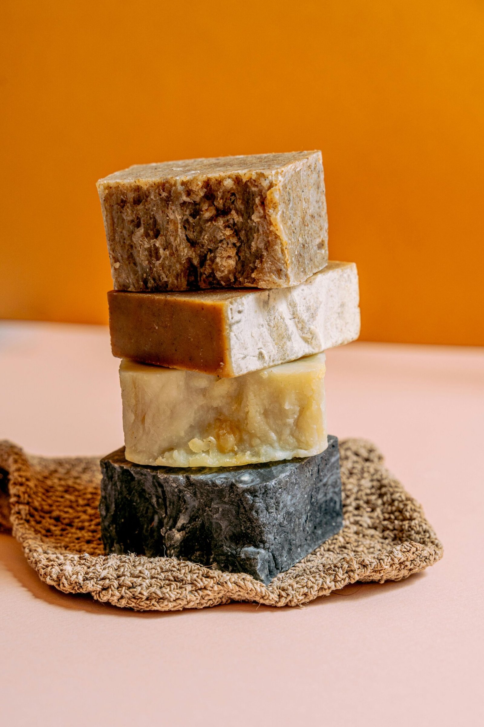 Four handmade natural soap bars stacked on a textured mat against a warm background.